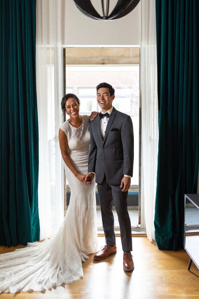 A groom, dressed in a Michael Kors Steel Grey Sterling, and a bride, wearing her wedding gown, standing in front of a window.