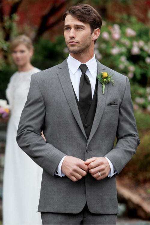 A man in an Allure Charcoal suit rental is standing next to a flower garden.