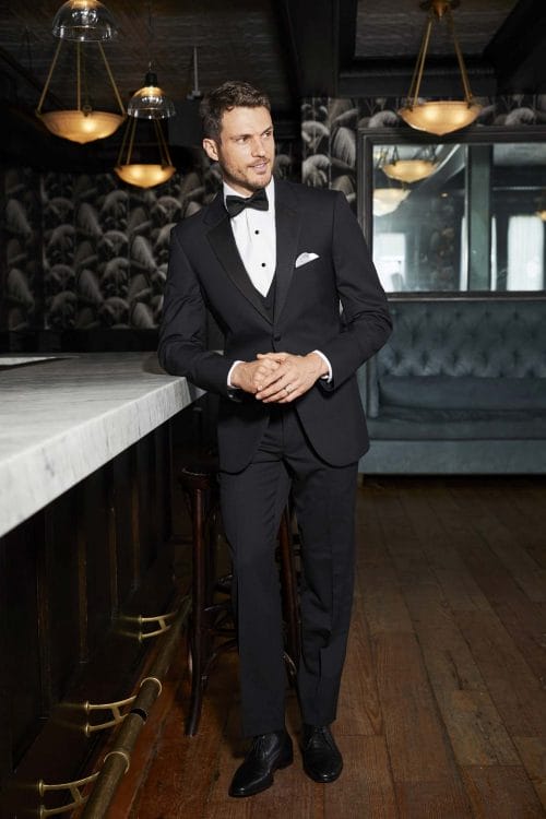 A man in an Allure Charcoal tuxedo rental standing in a bar.