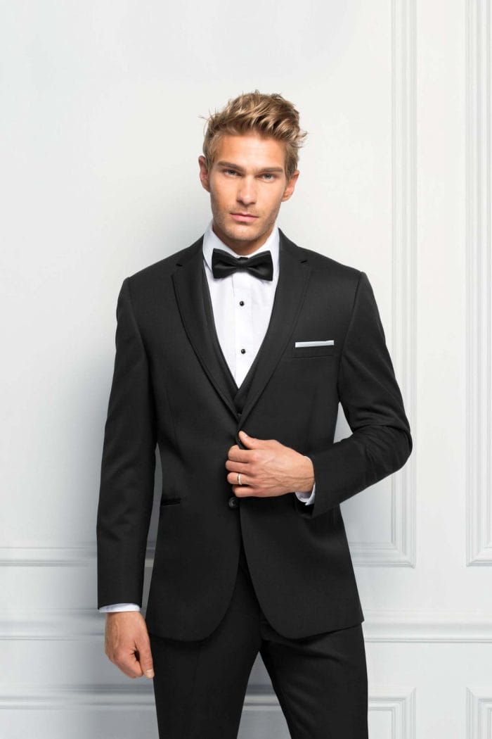 A man in an Allure Charcoal tuxedo posing for a photo at a suit rental shop.