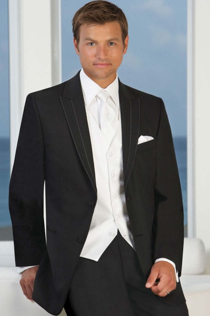 A man in an Allure Charcoal tuxedo is posing for a photo at a tuxedo rental shop.