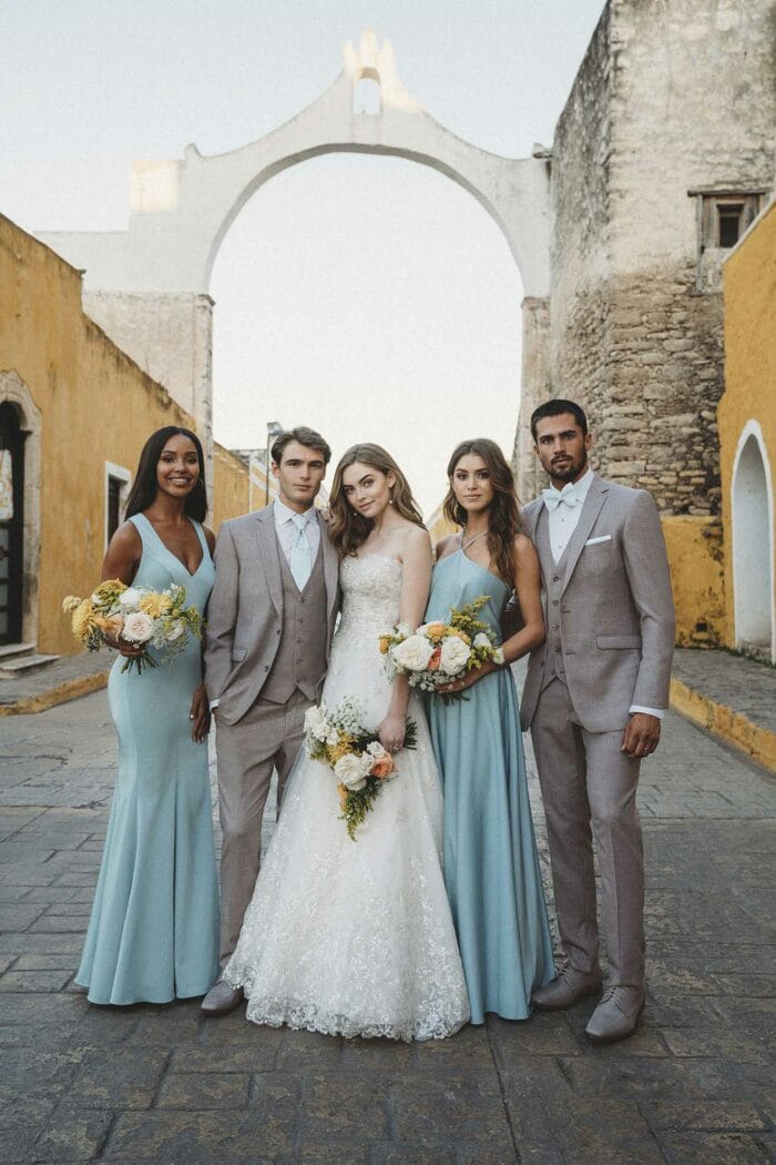 A group of bridesmaids posing in front of an archway, dressed in trendy Allure Heather Grey suits from a suit rental service.