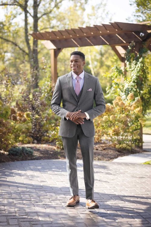 A man in a Michael Kors Medium Grey Performance suit rental standing in a park.