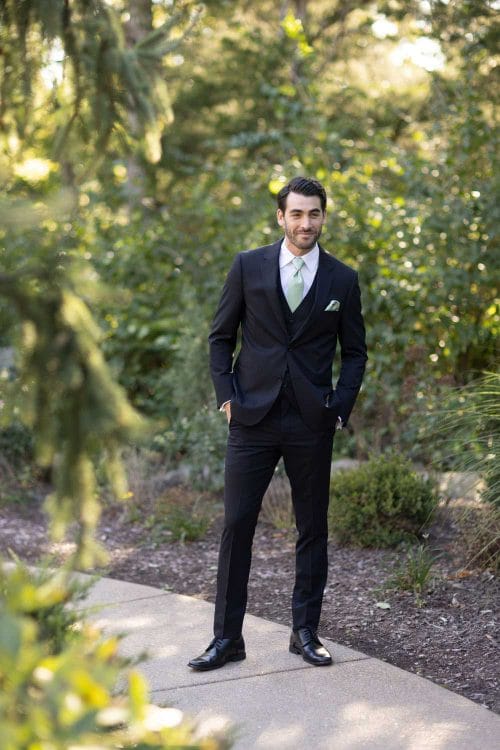 A groom in a Michael Kors Black Performance suit rental standing on a path with a green tie.